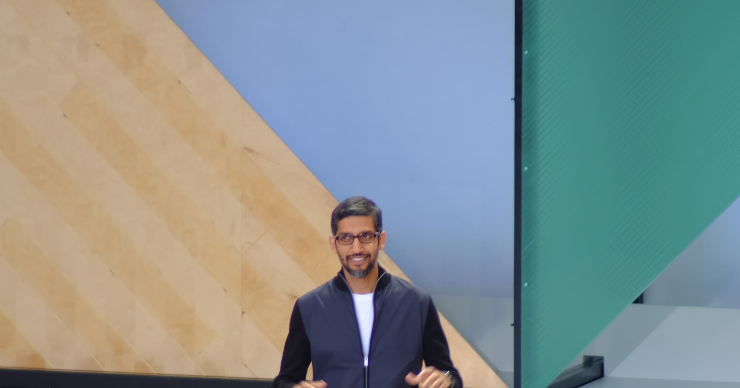 Google CEO Sundar Pichai gesturing on stage at Google I/O in a dark jacket over a white T-shirt