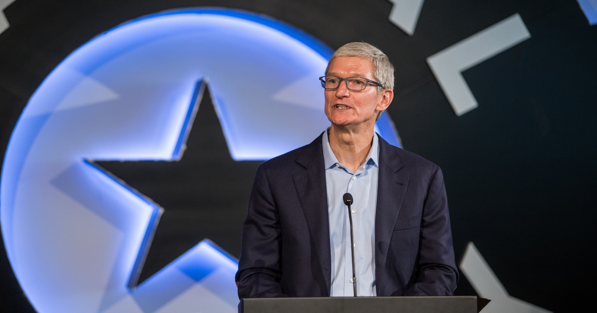 Apple CEO Tim Cook speaking at a podium in a navy blazer and light blue shirt, with a dark stage backdrop behind him