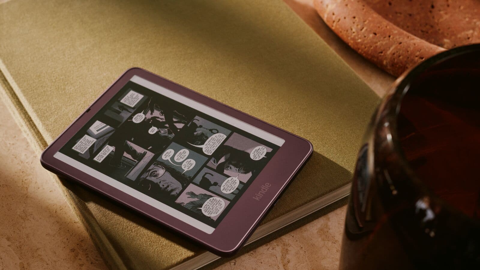 Purple Ube Amazon Kindle with a flush-front display resting on a green hardcover book, showing a comic page on the e-ink screen