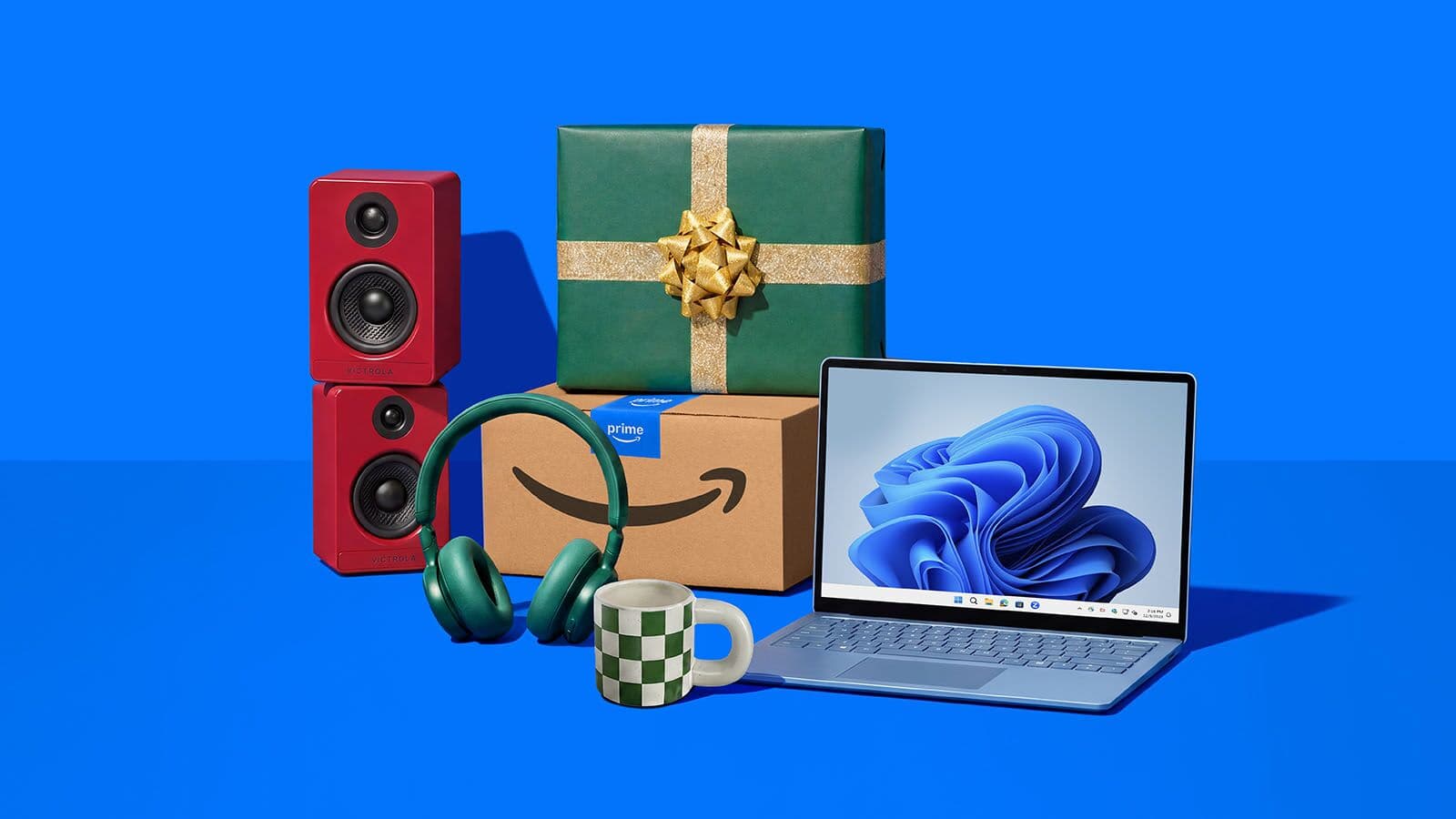 Official Amazon photo of a pair of red bookshelf speakers, green over-ear headphones, a wrapped green gift box sitting on an Amazon Prime box, a checkered mug and a silver laptop showing the Windows 11 bloom wallpaper on a blue background