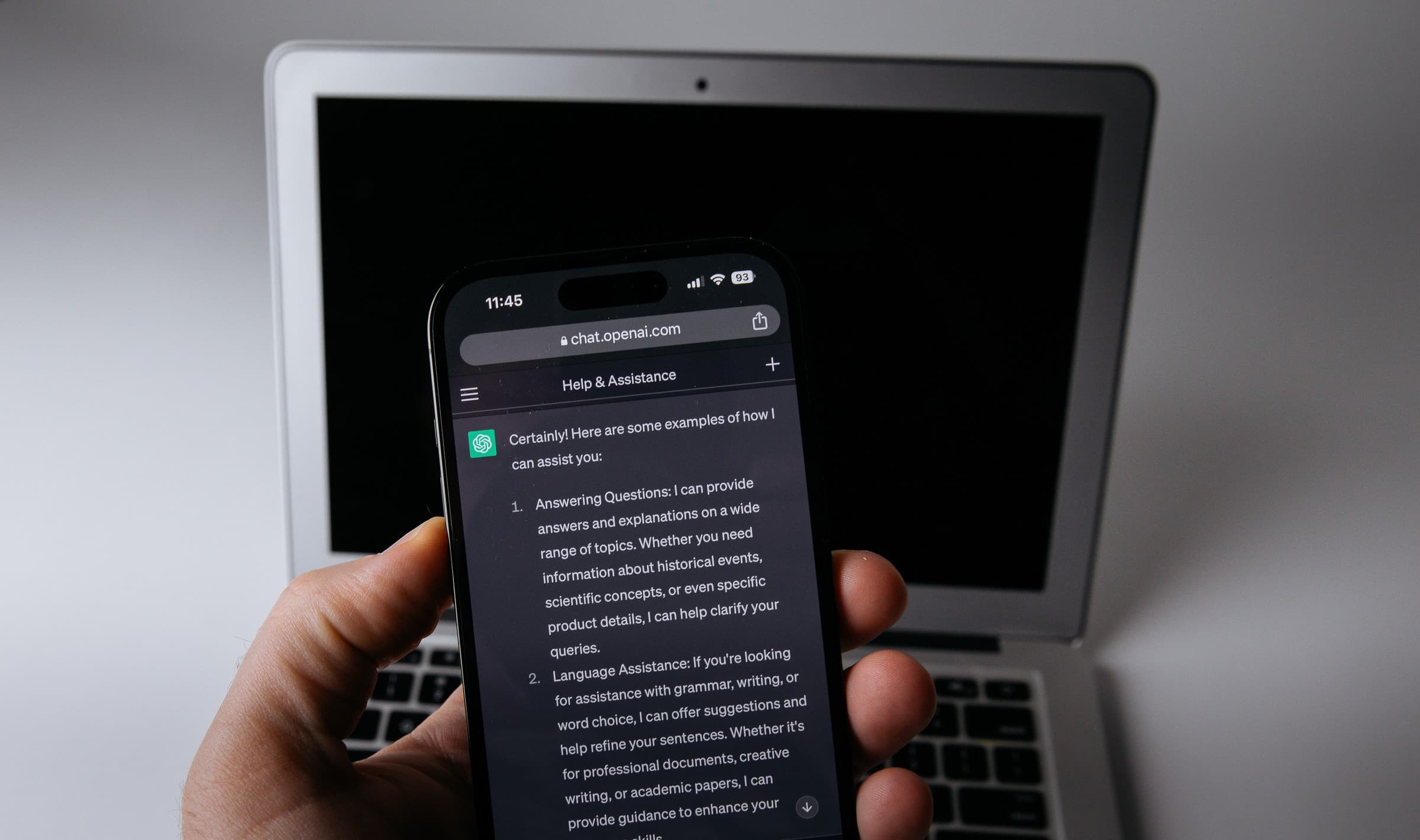 A hand holding a smartphone showing a ChatGPT reply on chat.openai.com that begins Certainly! Here are some examples of how I can assist you, with a silver laptop behind it