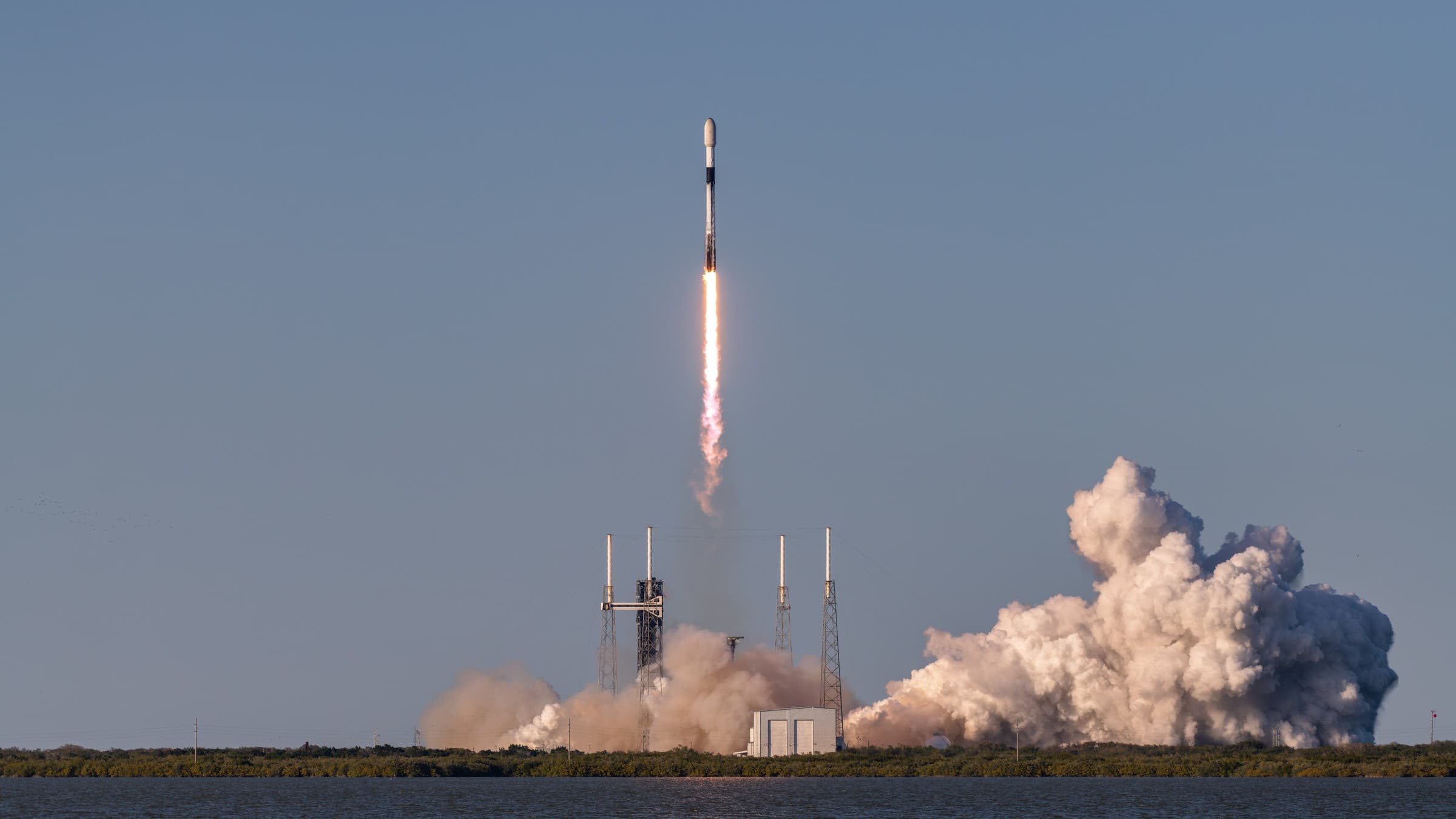 A SpaceX Falcon 9 rocket lifts off from Cape Canaveral on a column of flame carrying 24 Starlink satellites, with exhaust clouds billowing beside the launch pad and water in the foreground