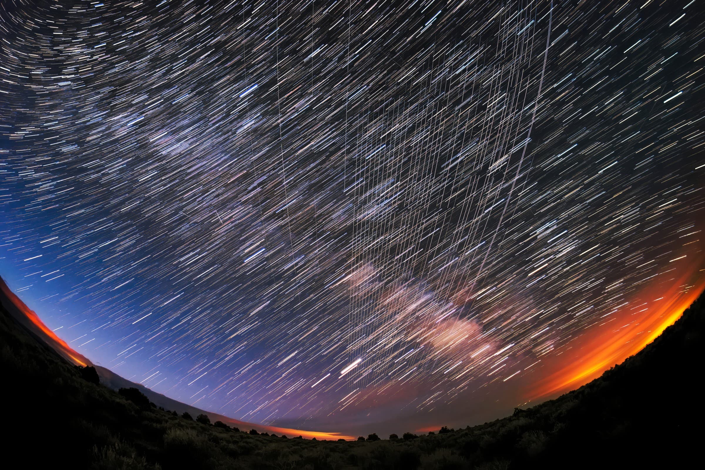 Long-exposure night photo over New Mexico in which a bundle of straight white Starlink satellite streaks cuts across curved star trails above a dark hillside and an orange glow on the horizon